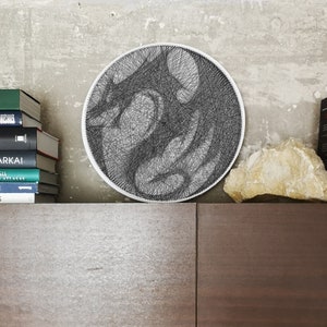 Puede incluir: Una pieza circular de arte de hilo que representa un dragón en hilo negro sobre un fondo blanco. La pieza de arte está colgada en una pared sobre un estante con libros y una roca.