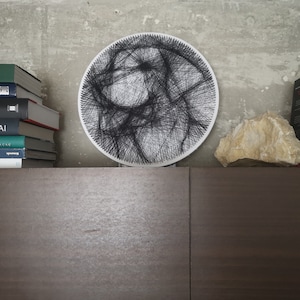 Puede incluir: Un marco circular de madera blanco con arte de cuerda negro en forma de cara. El marco está en un estante con una pila de libros y una roca.