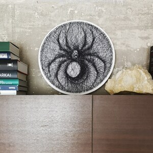 May include: A black and white string art spider design on a white circular canvas. The canvas is hanging on a wall with a stack of books and a rock below it.