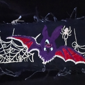 May include: A black fabric wrist cuff with a purple and red bat, white spider web, and a white spider embroidered on it. The cuff has a silver metal chain detail.