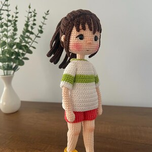 May include: A crocheted doll with brown hair, a white and green striped shirt, red shorts, and yellow shoes. The doll is standing on a wooden surface.