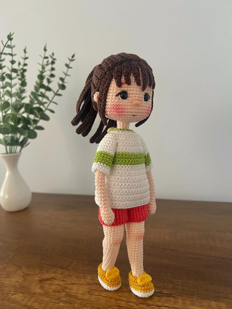 May include: A crocheted doll with brown hair, a white and green striped shirt, red shorts, and yellow shoes. The doll is standing on a wooden surface.