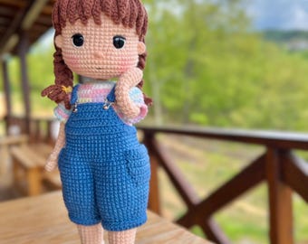 May include: A handmade crochet doll with brown hair in pigtails, wearing a light pink and blue striped shirt, blue overalls, and yellow shoes. The doll is standing on a wooden surface with a blurred green background.