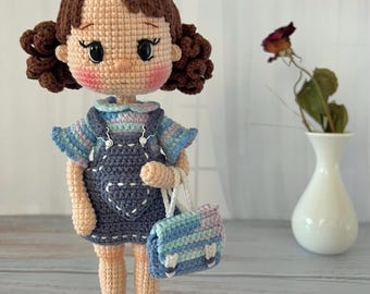 May include: A crocheted doll with brown hair, a blue and white striped shirt, a blue overall dress with a heart on the front, and blue and white striped shoes. The doll is holding a blue and white striped bag.