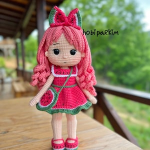 May include: A handmade crochet doll with a watermelon theme. The doll has pink hair, a red dress with a watermelon slice design, and a matching purse. The doll is wearing red shoes and a watermelon bow. The text "hobi parkim" is visible.