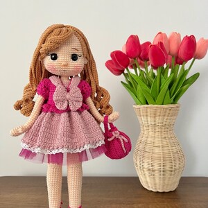 May include: A crocheted doll with brown hair, wearing a pink dress with a white lace trim and pink shoes. The doll is holding a pink crocheted purse. The doll is standing on a wooden surface in front of a vase of red tulips.