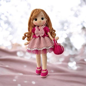 Puede incluir: Una muñeca de crochet con el pelo largo y castaño, que lleva un vestido rosa con encaje blanco y un lazo rosa. La muñeca lleva un bolso rosa de crochet.