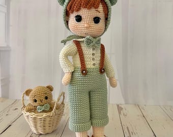 May include: A crocheted doll with brown hair, wearing a green hat with bear ears, a white shirt with a bow tie, green pants with suspenders, and brown shoes. The doll is standing next to a small crocheted bear in a wicker basket.