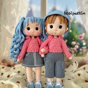 Peut inclure: Deux poupées crochetées à la main, un garçon et une fille, se tiennent côte à côte. La fille a des cheveux bleus et porte un sweat à capuche rose et une jupe grise. Le garçon a des cheveux bruns et porte un chapeau bleu, un sweat à capuche rose et un short gris. Les deux poupées ont des chaussures bleues.