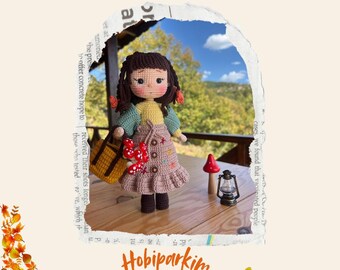 May include: Crochet pattern for a Defne doll, a handmade amigurumi doll with brown hair, a green sweater, a brown skirt, and a basket. The doll is sitting on a wooden table with a mushroom and a lantern. The image has the text "Pattern for Defne Doll" and "Hobiparkim SEPTEMBER 2024".