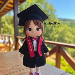 May include: A crocheted doll wearing a graduation cap, gown, and sash with "2025" on it. The doll has brown hair, rosy cheeks, and is wearing a white shirt and red bow tie. The doll is wearing white sneakers with red accents.