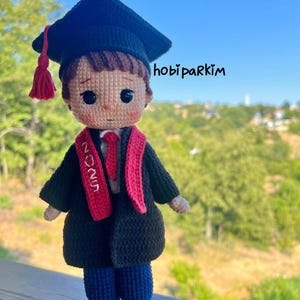 May include: A handmade crochet doll dressed in a graduation outfit. The doll wears a black cap with a red tassel, a black gown, and a red sash with "2025" on it. The doll has blue pants, white shoes with red laces, and the text "hobi parkim".