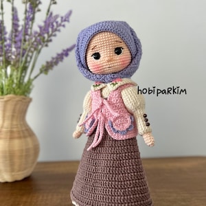 Op de afbeelding: Een handgemaakte gehaakte pop met een lavendelkleurige hoofddoek, een roze vest en een bruine rok. De pop heeft roze wangen en staat op een houten oppervlak. De tekst "hobi parkim" is zichtbaar aan de rechterkant van de pop.