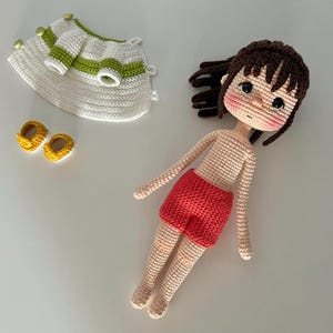 May include: A crocheted doll with brown hair, wearing a red skirt and a white crocheted dress with green stripes. The doll is laying on a white surface with a pair of yellow crocheted shoes.