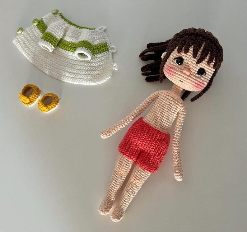May include: A crocheted doll with brown hair, wearing a red skirt and a white crocheted dress with green stripes. The doll is laying on a white surface with a pair of yellow crocheted shoes.