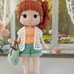 May include: A handmade crochet doll with red hair in a ponytail. The doll is dressed in a white open-weave cardigan, an orange top, and teal shorts. She carries a teal handbag and wears orange and teal sandals. The doll is approximately 25 cm tall.