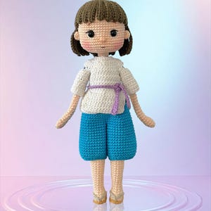 May include: A handmade crochet doll with brown hair and rosy cheeks. The doll wears a white top with a purple belt, blue shorts, and beige shoes. The doll is approximately 8 inches tall, a charming handcrafted item.