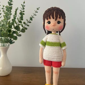 May include: A crocheted doll with brown hair, a white shirt with a green stripe, red shorts, and yellow shoes.