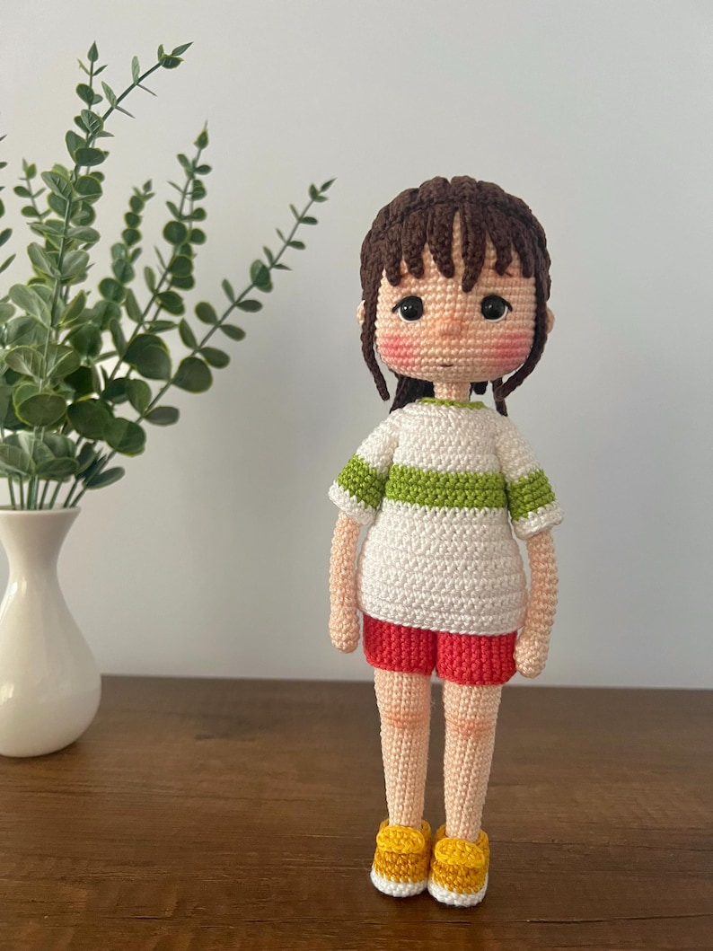 May include: A crocheted doll with brown hair, a white shirt with a green stripe, red shorts, and yellow shoes.