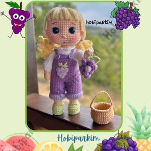May include: A crochet doll pattern with the text "Duru Doll Crochet Pattern." The doll has blonde hair, a purple outfit, and is holding a bunch of grapes. A small basket and grape illustrations are also featured. The pattern is from Hobiparkim.