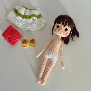 May include: A crocheted doll with brown hair, wearing a white dress, red shorts, and yellow shoes. The doll is laying on a white surface.