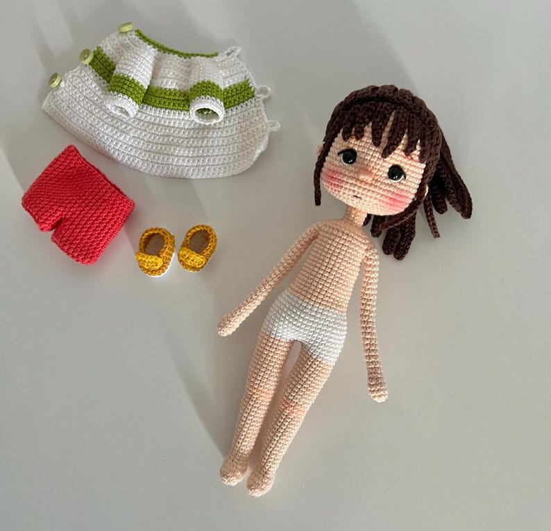 May include: A crocheted doll with brown hair, wearing a white dress, red shorts, and yellow shoes. The doll is laying on a white surface.