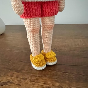 May include: A crocheted doll wearing a white shirt, red shorts, and yellow shoes. The doll is standing on a wooden surface.
