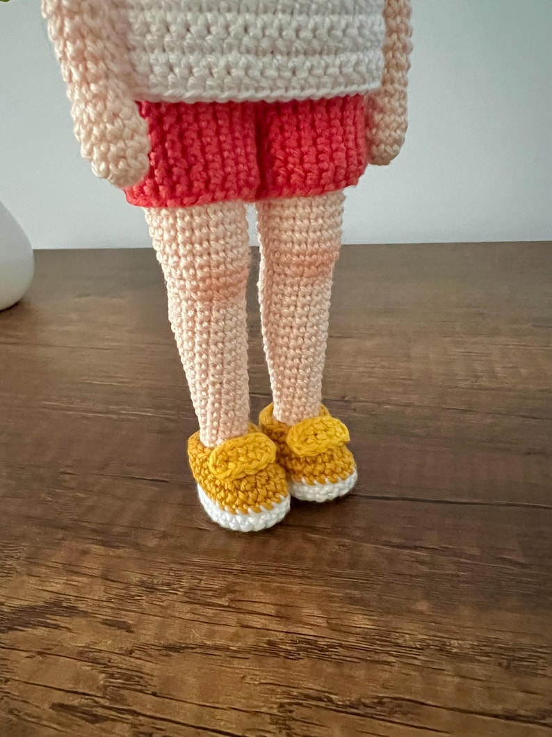 May include: A crocheted doll wearing a white shirt, red shorts, and yellow shoes. The doll is standing on a wooden surface.