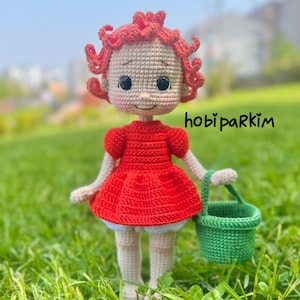 May include: A handmade crochet doll with red hair and a red dress, holding a green basket. The doll has a beige face with black eyes and is standing on green grass. The text "hobi parkim" is visible on the right side.