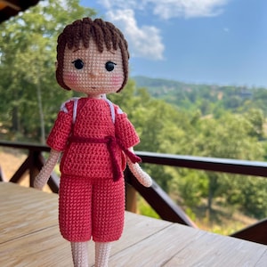 May include: A handmade crochet doll featuring a character from a popular animated film. The doll is dressed in a red outfit with white accents and a matching red belt. The doll has brown hair and is standing on a wooden surface.