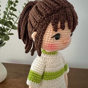 May include: A crocheted doll with brown hair in a ponytail, wearing a white and green striped shirt. The doll has a pink blush on its cheeks and black eyes.