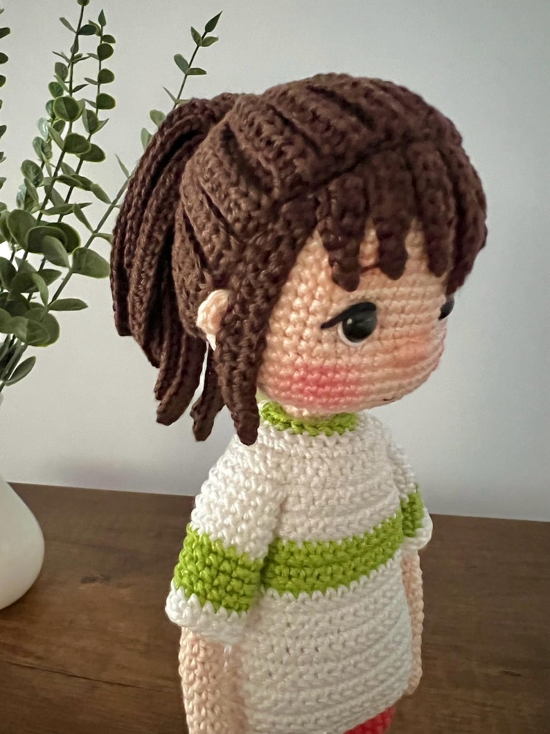 May include: A crocheted doll with brown hair in a ponytail, wearing a white and green striped shirt. The doll has a pink blush on its cheeks and black eyes.