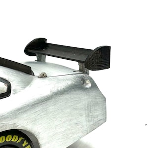 Może przedstawiać: Srebrny samochód wyścigowy z czarnym tylnym spojlerem. Samochód ma czarną oponę z napisem "GOODYEAR" na niej.
