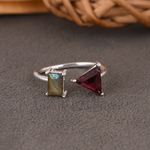 May include: A silver ring featuring a rectangular labradorite gemstone and a triangular garnet gemstone. The ring is open on one side and has a delicate design.