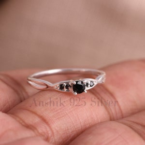 May include: A silver ring featuring a central round black stone flanked by two smaller black stones on each side. The band has a split design that converges at the setting. The text Anshik 925 Silver is visible across the lower portion of the ring.