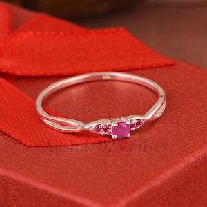 Puede incluir: Un anillo de plata con una banda delicada y retorcida y una pequeña piedra preciosa rosa en el centro. El anillo está engastado con piedras preciosas rosas más pequeñas a cada lado de la piedra central.