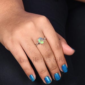 K&ouml;nnte beinhalten: Ein silberner Ring mit einem ovalen Opal-Edelstein. Der Opal zeigt eine Mischung aus Gr&uuml;n, Gelb und Blau. Das Ringband hat ein dekoratives, wirbelndes Design. Der Text "Ansik 925 Silver" ist im Bild zu sehen.