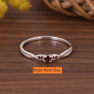 May include: A silver ring with a red gemstone and smaller red gemstones on the band. The ring is on a wooden surface. Ships Next Day.