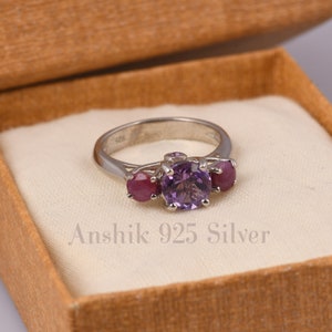 May include: A silver ring featuring a large purple amethyst gemstone and two smaller red ruby gemstones. The ring is displayed in a white box with the inscription "Anshik 925 Silver".