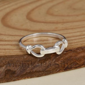 Puede incluir: Un anillo de plata pulida que presenta un diseño central de barra horizontal conectado a dos extremos en forma de bucle. La banda es lisa y circular, mostrando un acabado metálico que refleja la luz a través de toda su superficie.
