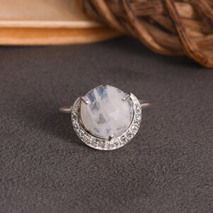 May include: A silver ring featuring a large, round, iridescent moonstone center stone. The moonstone is surrounded by a halo of small, clear gemstones. The ring is set on a dark gray surface.