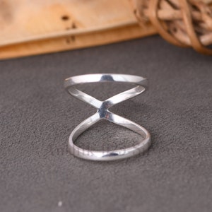 May include: A close-up of a silver ring with an X-shaped design. The ring has a polished finish and a minimalist style. The ring is on a gray surface.
