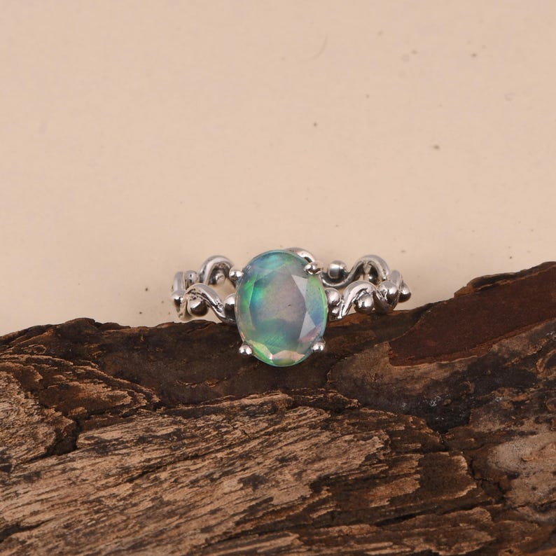 K&ouml;nnte beinhalten: Ein silberner Ring mit einem ovalen Opal-Edelstein. Der Opal hat eine Mischung aus Gr&uuml;n, Blau und Lila. Das Band des Rings weist ein dekoratives, wirbelndes Design auf. Der Ring wird auf einem Holzst&uuml;ck pr&auml;sentiert.