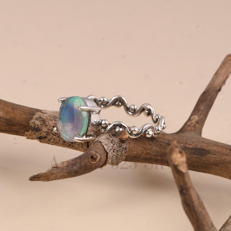 K&ouml;nnte beinhalten: Ein silberner Ring mit einem ovalen, irisierenden Edelstein. Das Band hat ein dekoratives, wellenf&ouml;rmiges Design. Der Ring wird auf einem Holzst&uuml;ck vor einem neutralen Hintergrund pr&auml;sentiert. Der Edelstein hat Schattierungen von Blau, Gr&uuml;n und Lila.