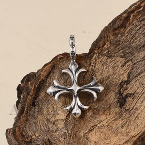 Puede incluir: Colgante de plata con un diseño de cruz estilizada. La cruz presenta cuatro brazos curvos con un punto central y una argolla decorativa. El colgante se muestra sobre un fondo de madera texturizada.