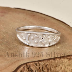 May include: A silver ring with a celestial design featuring a crescent moon, stars, and other shapes. The ring is set against a wooden surface. The text "Anshik 925 Silver" is visible at the bottom of the image.