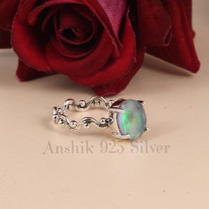 K&ouml;nnte beinhalten: Ein silberner Ring mit einem ovalen, schillernden Edelstein. Das Band hat ein zartes, wellenf&ouml;rmiges Design mit kleinen, runden Akzenten. Der Text "Anshik 925 Silver" ist darunter zu sehen. Eine dunkelrote Rose ist im Hintergrund.