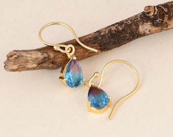 May include: A pair of gold-colored drop earrings. Each earring features a teardrop-shaped, bi-color gemstone with hues of blue and purple. The gemstones are set in a gold-colored bezel and suspended from a curved ear wire.