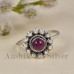 May include: A sterling silver ring featuring a central round cabochon-cut deep pink gemstone. The stone is surrounded by a twisted rope border and a halo of small silver spheres. The text Anshik Silver is visible at the bottom of the image.