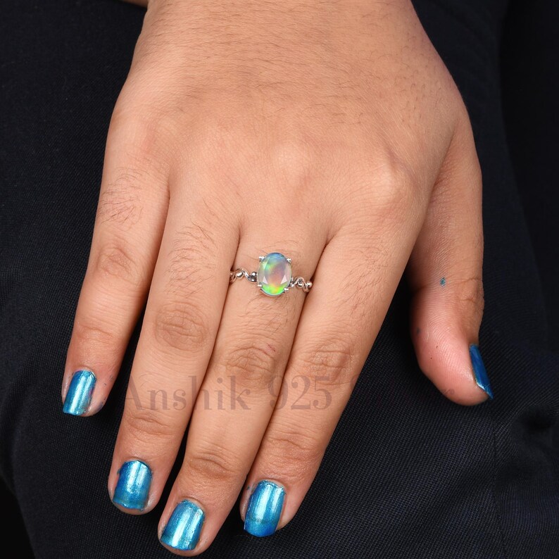 K&ouml;nnte beinhalten: Ein silberner Ring mit einem ovalen Opal-Edelstein. Der Opal zeigt eine Mischung aus Farben, darunter Gr&uuml;n, Blau und Gelb. Das Ringband hat ein filigranes, verziertes Design. Der Text "Anshik 925" ist sichtbar.